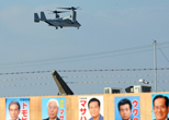 金武町議会議員選挙ポスター掲示板の向こうに見えるオスプレイ。2012年11月